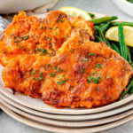 Two fried pork chops on a stack of small round plates. Pork chops are plated with green beans and a lemon wedge.