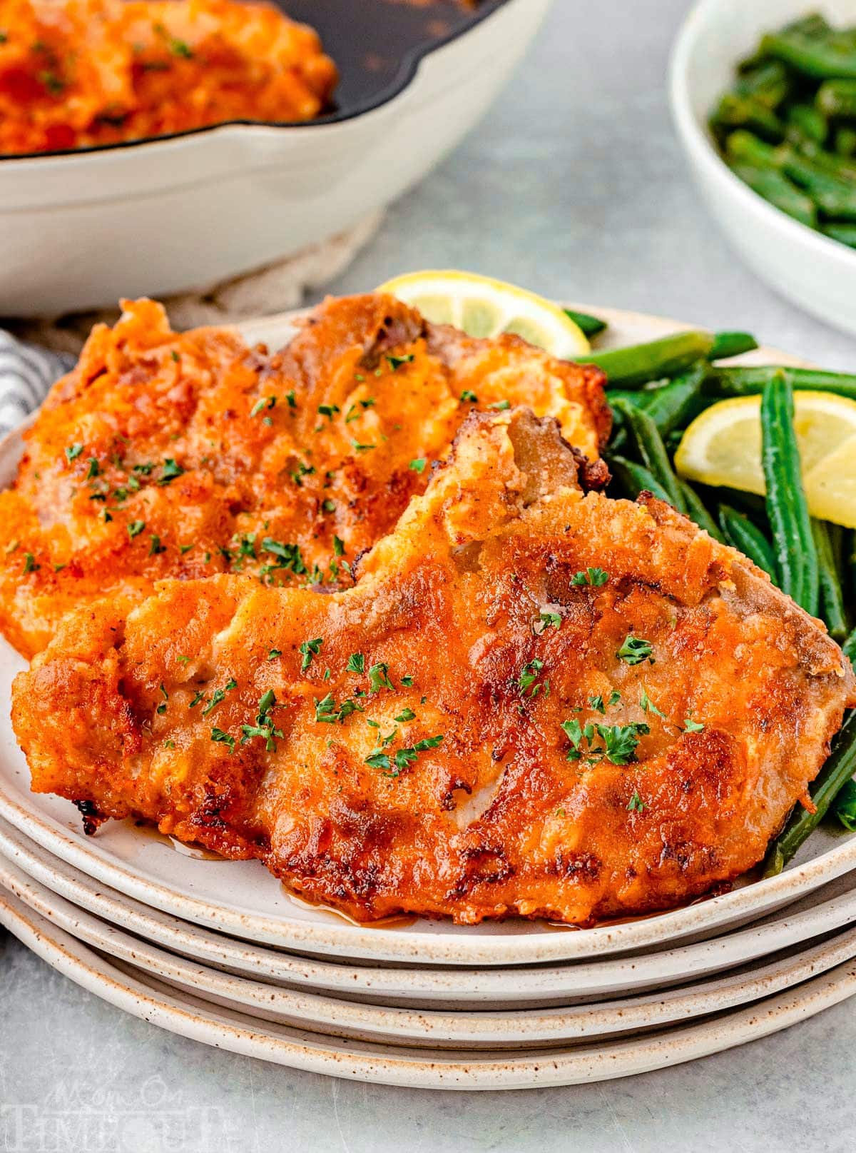 Two fried pork chops on a stack of small round plates. Pork chops are plated with green beans and a lemon wedge.