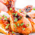 Honey glazed chicken drumstick garnished with green onions being picked up off a small sheet tray with more chicken on it.