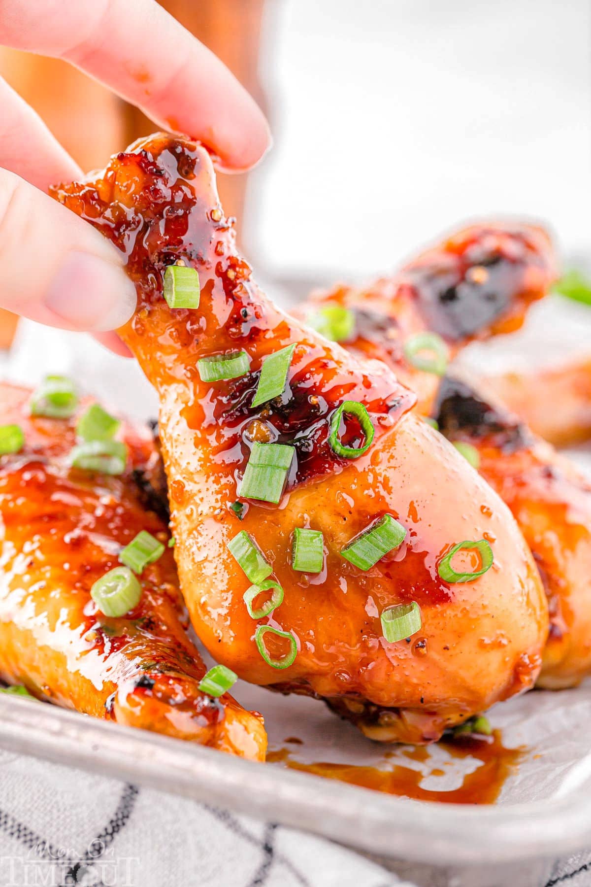 Honey glazed chicken drumstick garnished with green onions being picked up off a small sheet tray with more chicken on it.