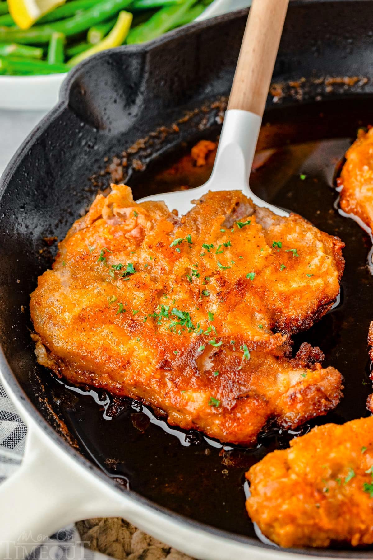 A spatula holding a fried pork chop in a cast iron skilet.