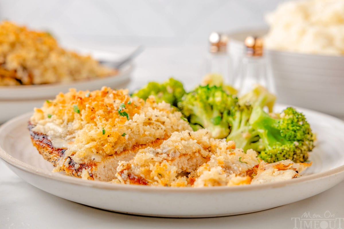 Wide shot of longhorn parmesan crusted chicken recipe on plate with broccoli on the backside of the plate. More chicken and mashed potatoes can be seen in the background.