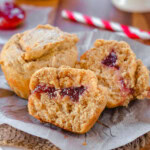 Two peanut butter and jelly muffins on a piece of parchment. One muffin has been split in half to reveal the jelly center.