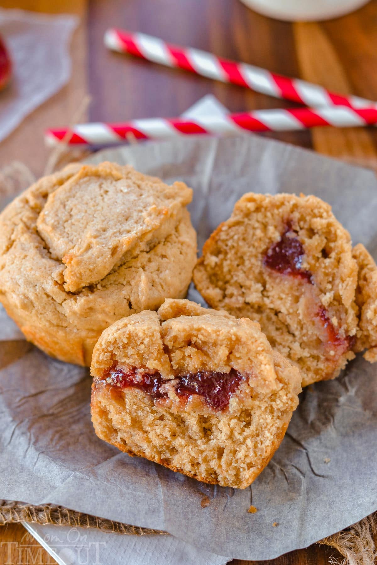 Two peanut butter and jelly muffins on a piece of parchment. One muffin has been split in half to reveal the jelly center.