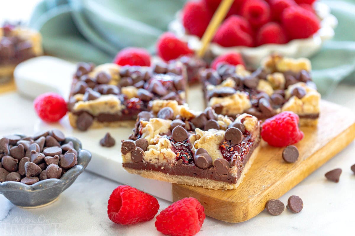 Wide shot of three bars sitting on a small white marble and wood cutting board next to each other with chocolate chips and raspberries sprinkled around them.