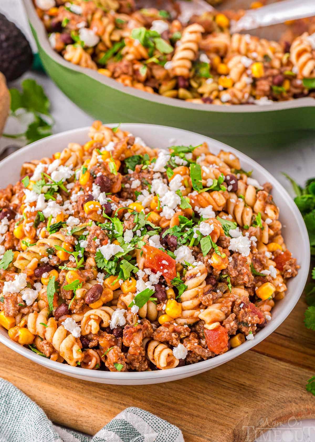 angled view of bowl filled with taco pasta and green skillet in the background with the rest of the taco pasta recipe. 