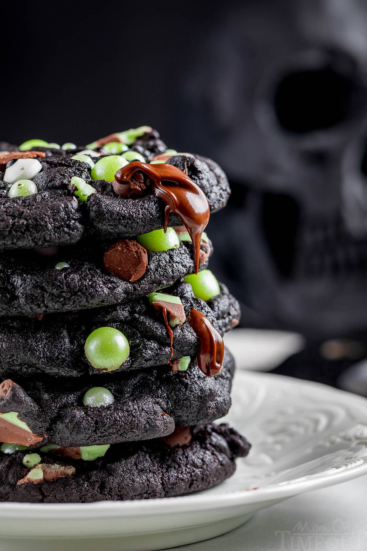 Side shot of stack of halloween cookies made with black cocoa powder stacked on a white plate with melty chocolate chips and green pearl candies.