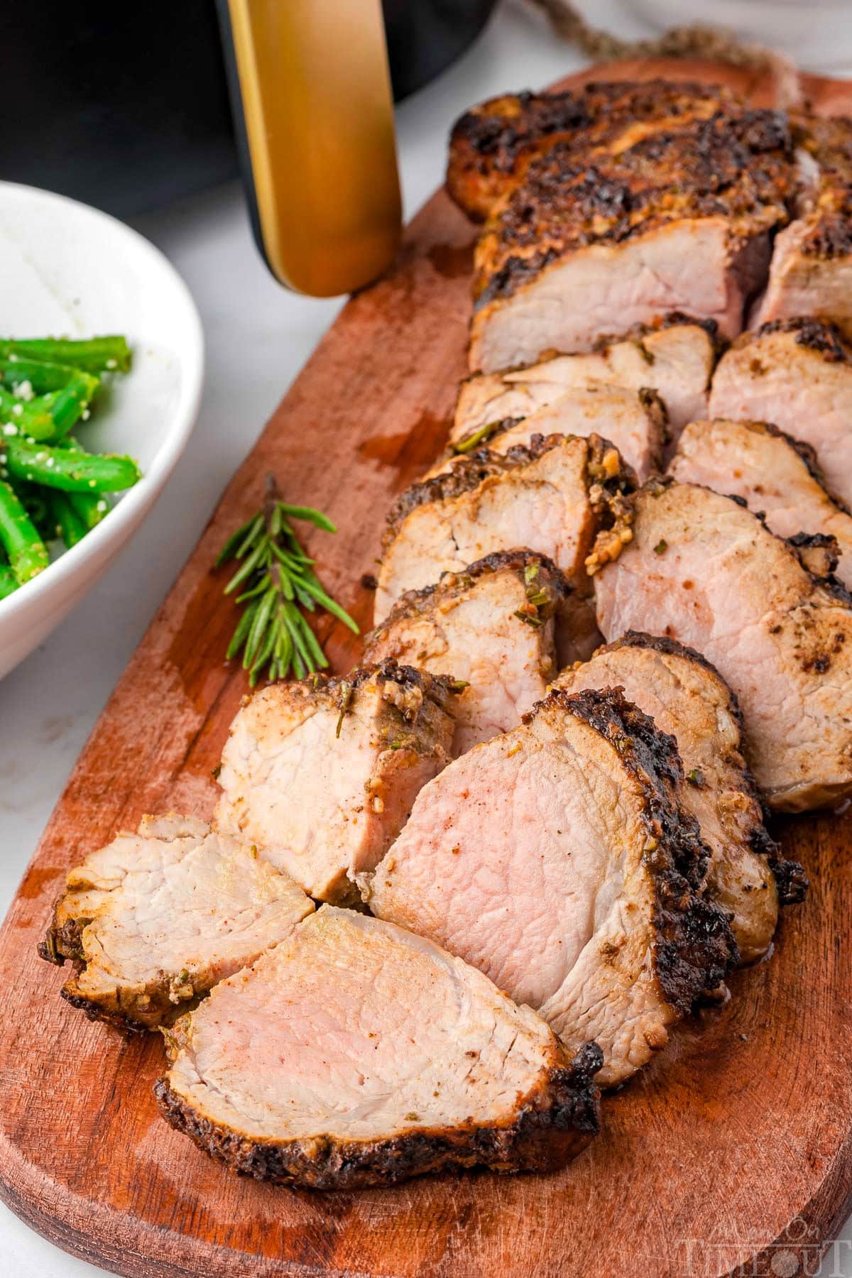 Air fryer pork tenderloin cut into pieces on a wood cutting board with air fryer in background.