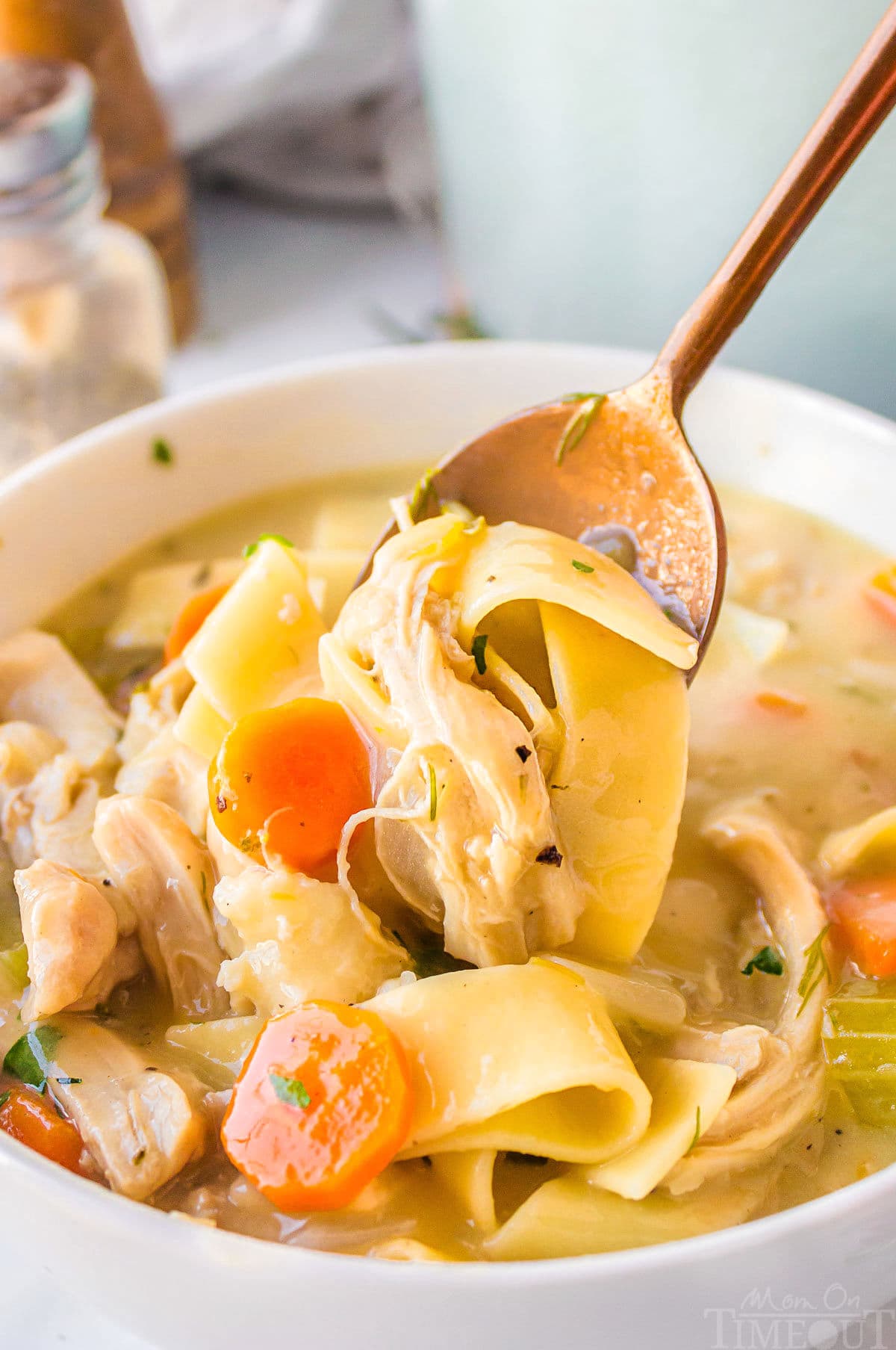 A spoon holding chicken and noodles over a bowl of chicken noodle soup.