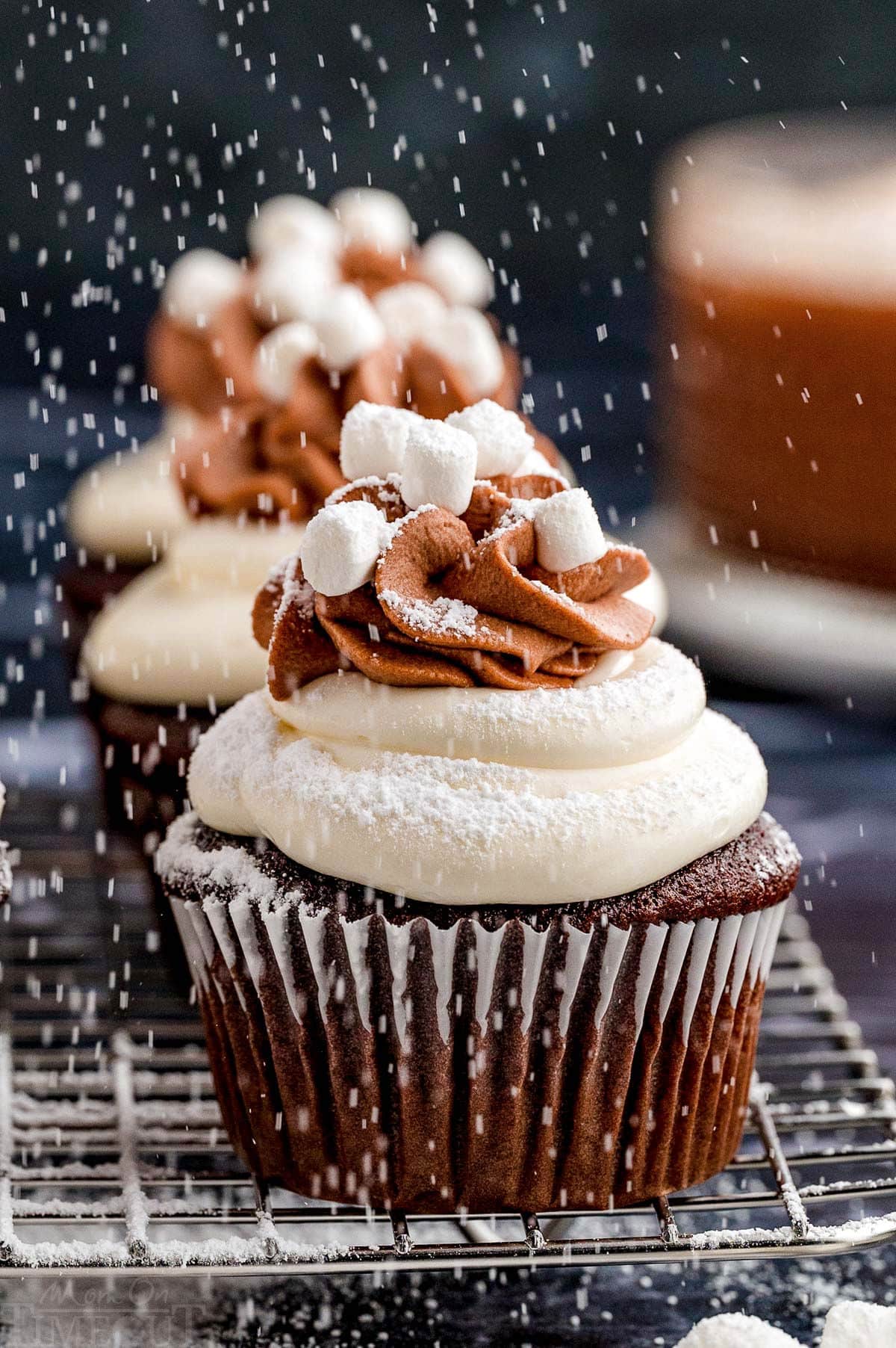 Hot chocolate cupcakes on a metal cooling rack. Powdered sugar is dusting the cupcakes from above.