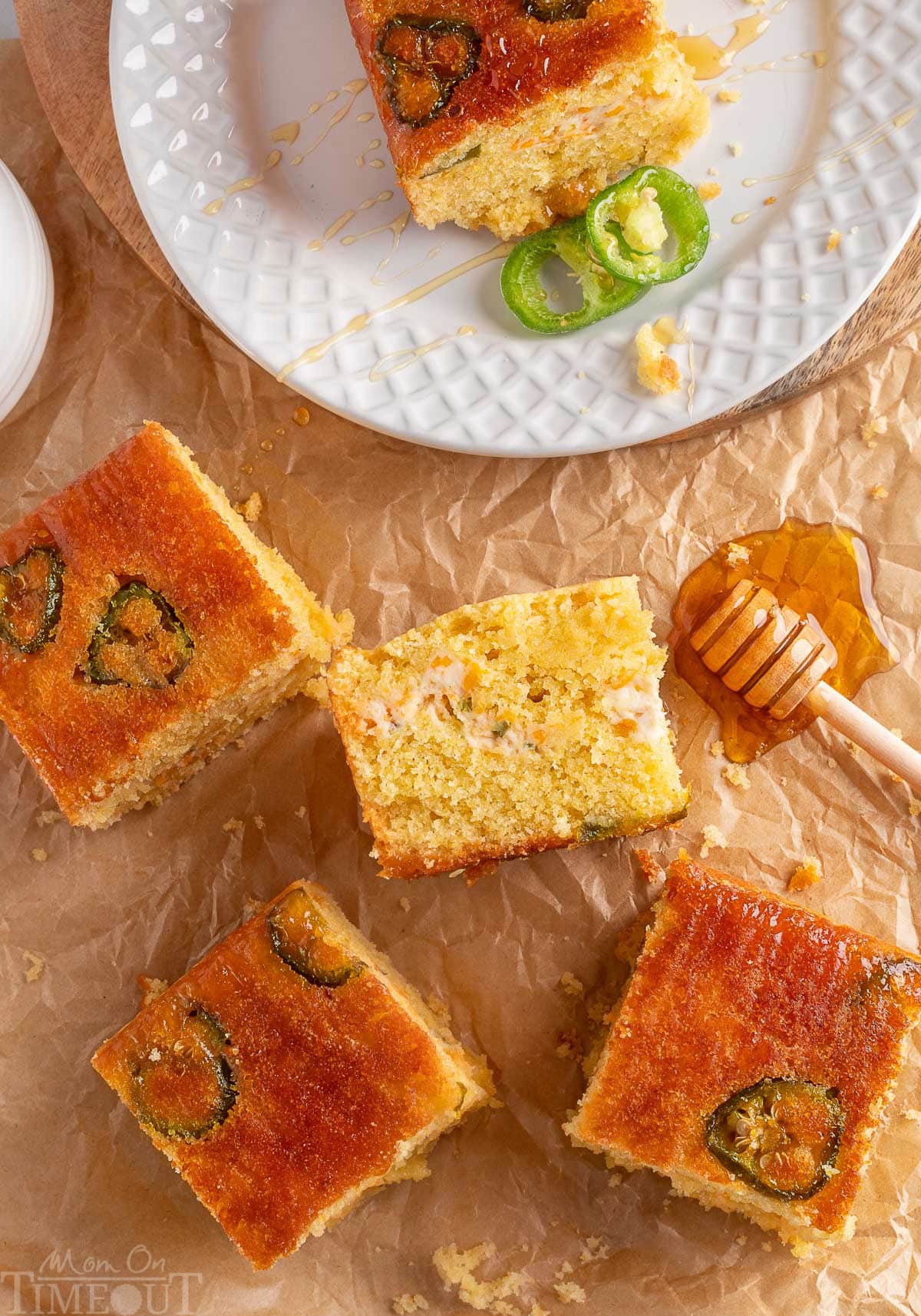 Jalapeno cornbread cut into squares on brown parchment paper. One piece is on a white plate with two slices of jalapeno next to it.