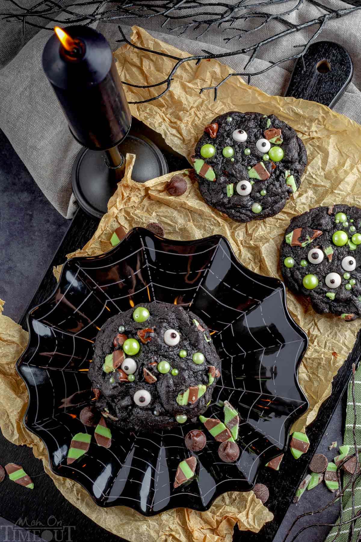 Halloween monster cookies on a black plate with a spider web design. Two cookies are on a piece of brown parchment paper. A black candle and black tree branch are next to them.