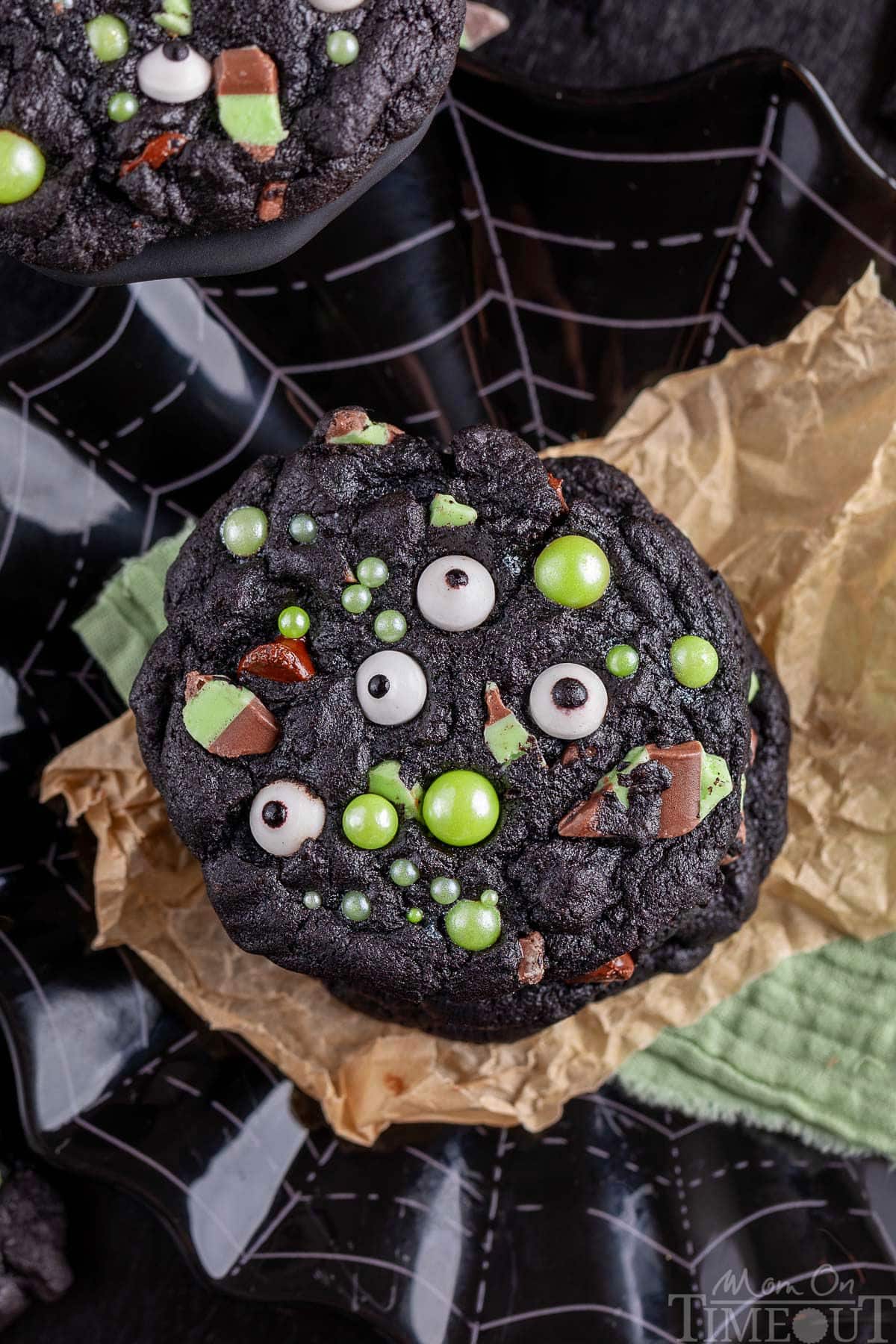 Monster Halloween Cookies stacked on brown parchment and black web design plate. Cookies have candy eyes and green sugar pearls for decoration.