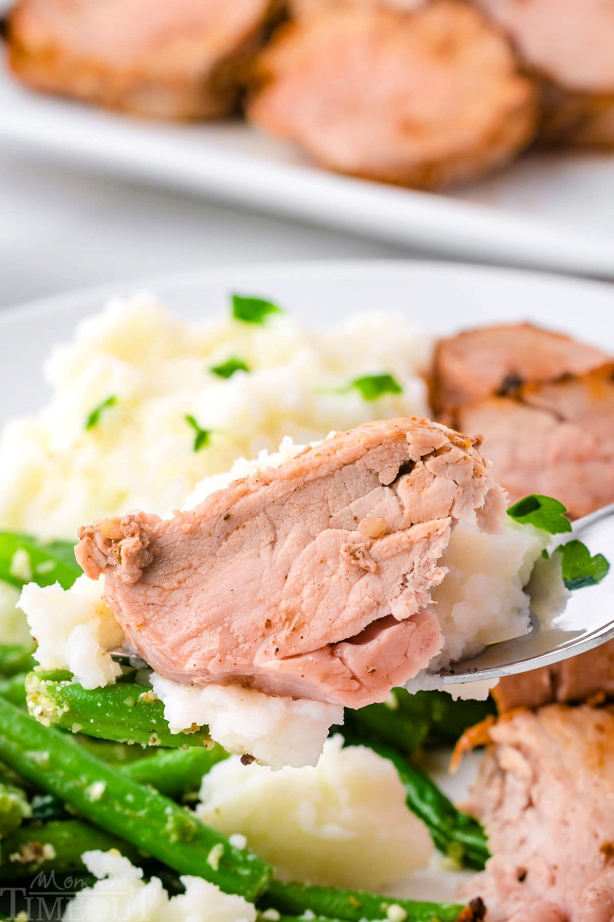 Forkful of pork tenderloin being lifted up from a plate with mashed potatoes and green beans.