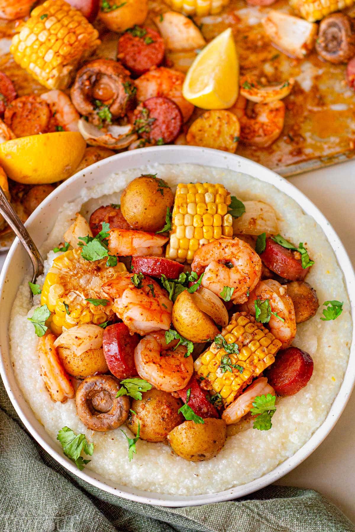 Shrimp boil in a bowl served with grits. The bowl is sitting next to a sheet pan with the rest of the shrimp boil.