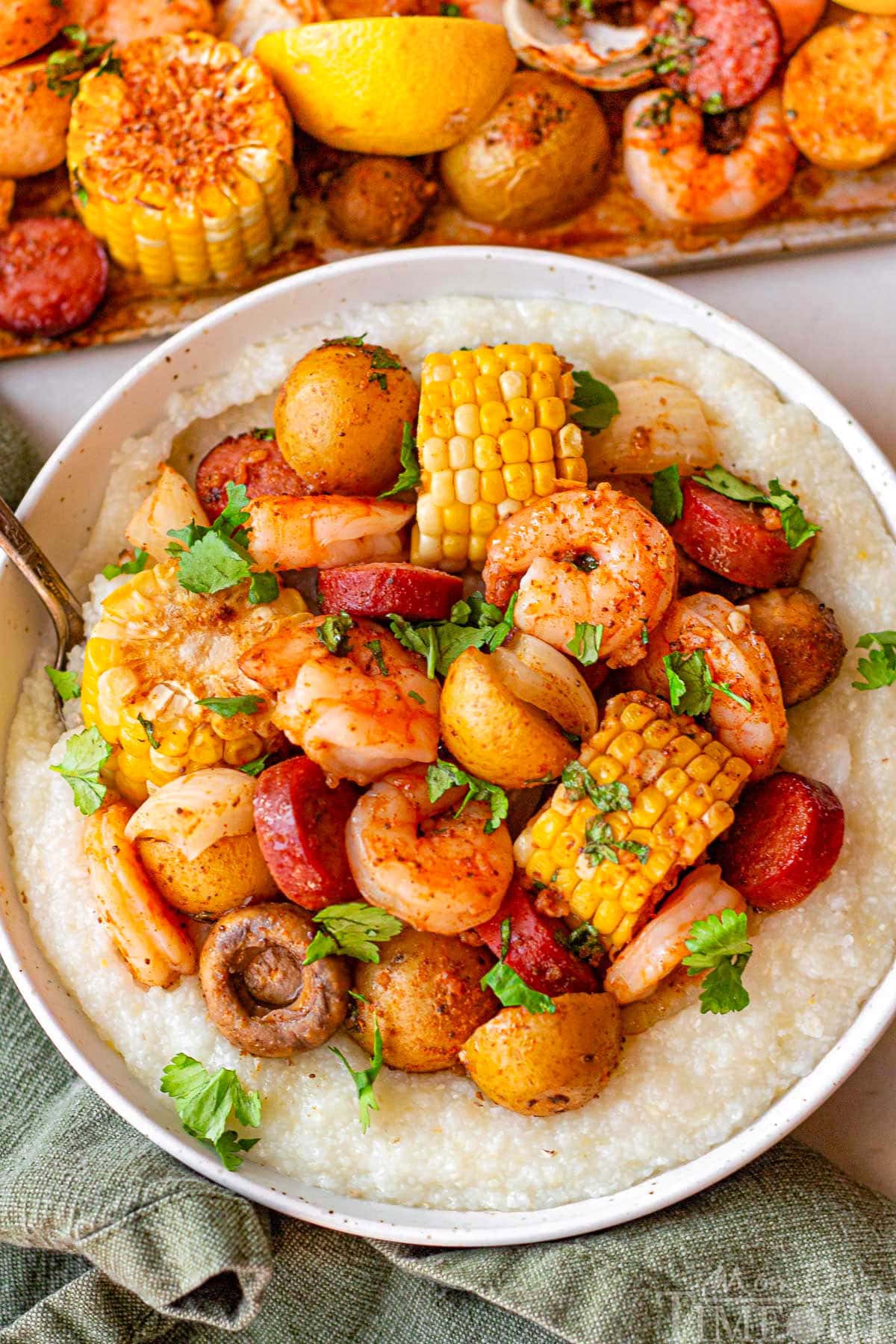 Shrimp boil in a bowl served with grits. The bowl is sitting next to a sheet pan with the rest of the shrimp boil.