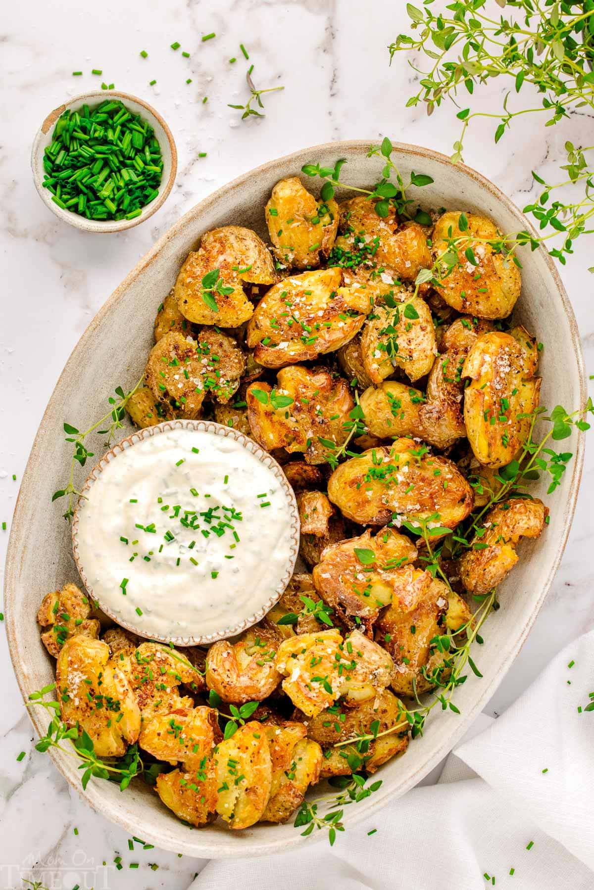 Top down view of an oval platter of smashed potatoes garnished with thyme and chives. A small bowl of dip is nestled amongst the smashed potatoes.