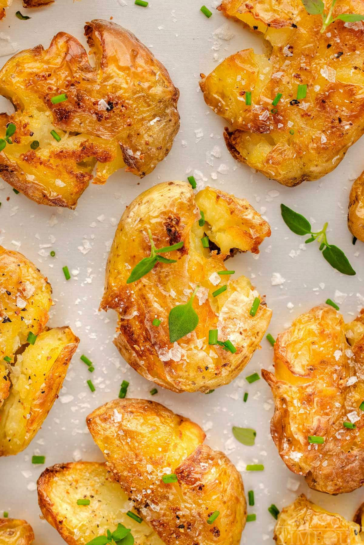 Top down view of smashed potatoes recipe on a white surface. The potatoes have been seasoned with salt, pepper and are topped with chives and thyme.