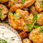 Close up of smashed potatoes on a plate garnished with chives and thyme sitting next to a small bowl of dip.