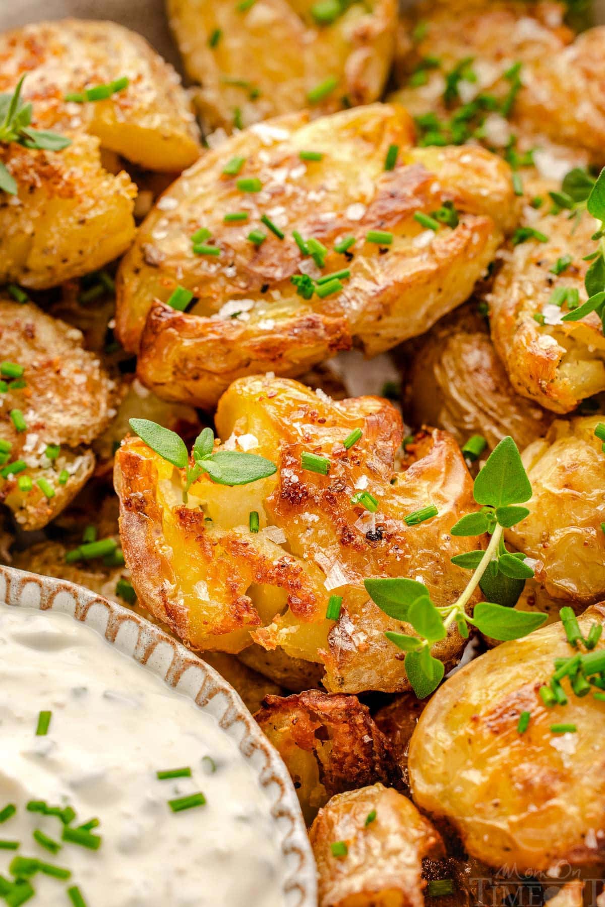 Close up of smashed potatoes on a plate garnished with chives and thyme sitting next to a small bowl of dip.