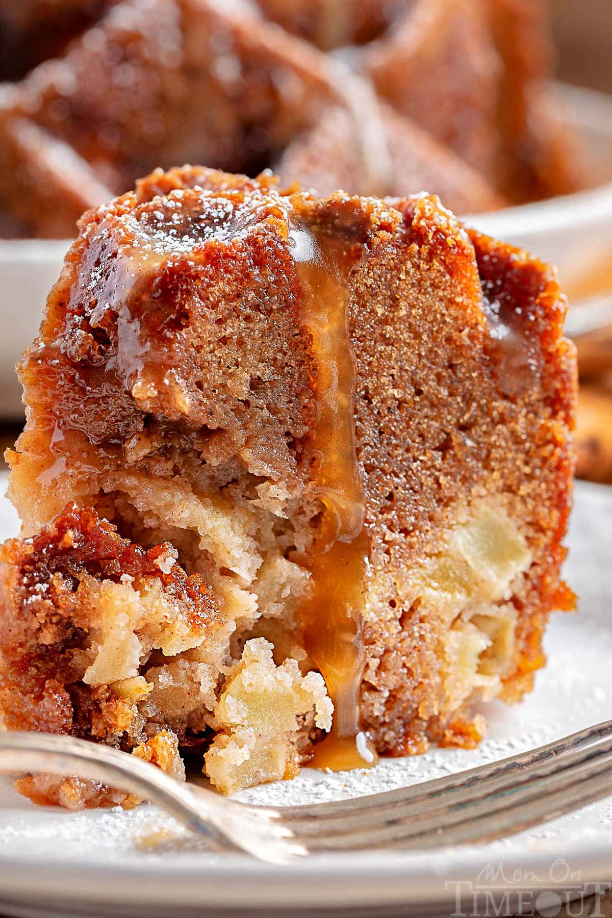 Close look at piece of caramel apple bundt cake topped with a caramel glaze sitting on a white round plate. The rest of the apple bundt cake can be seen in the background.