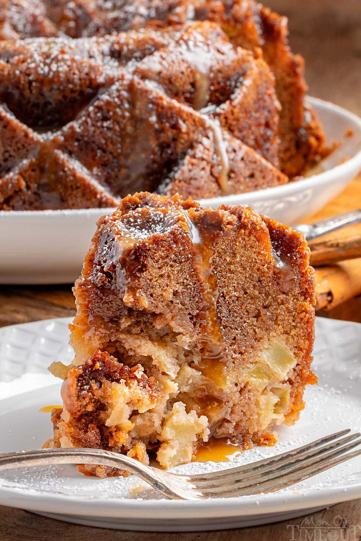 Piece of apple bundt cake topped with a caramel glaze sitting on a white round plate. The rest of the apple bundt cake can be seen in the background.