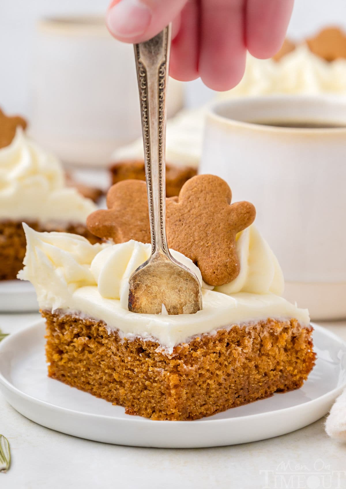 A hand putting a fork into a slice of gingerbread cake. The cake is on a white plate, and mugs of coffee and another piece of cake are in the background.