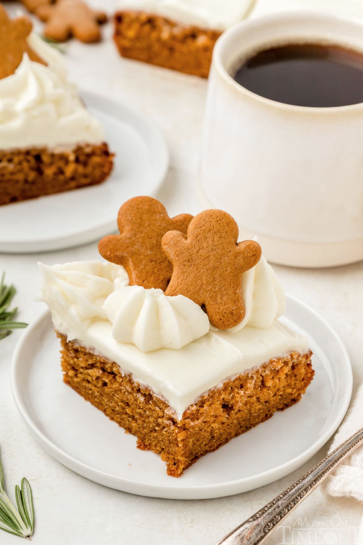 Angled view looking down on a piece of gingerbread cake topped with cream cheese frosting on a round white plate. A cup of coffee can be seen in the background.