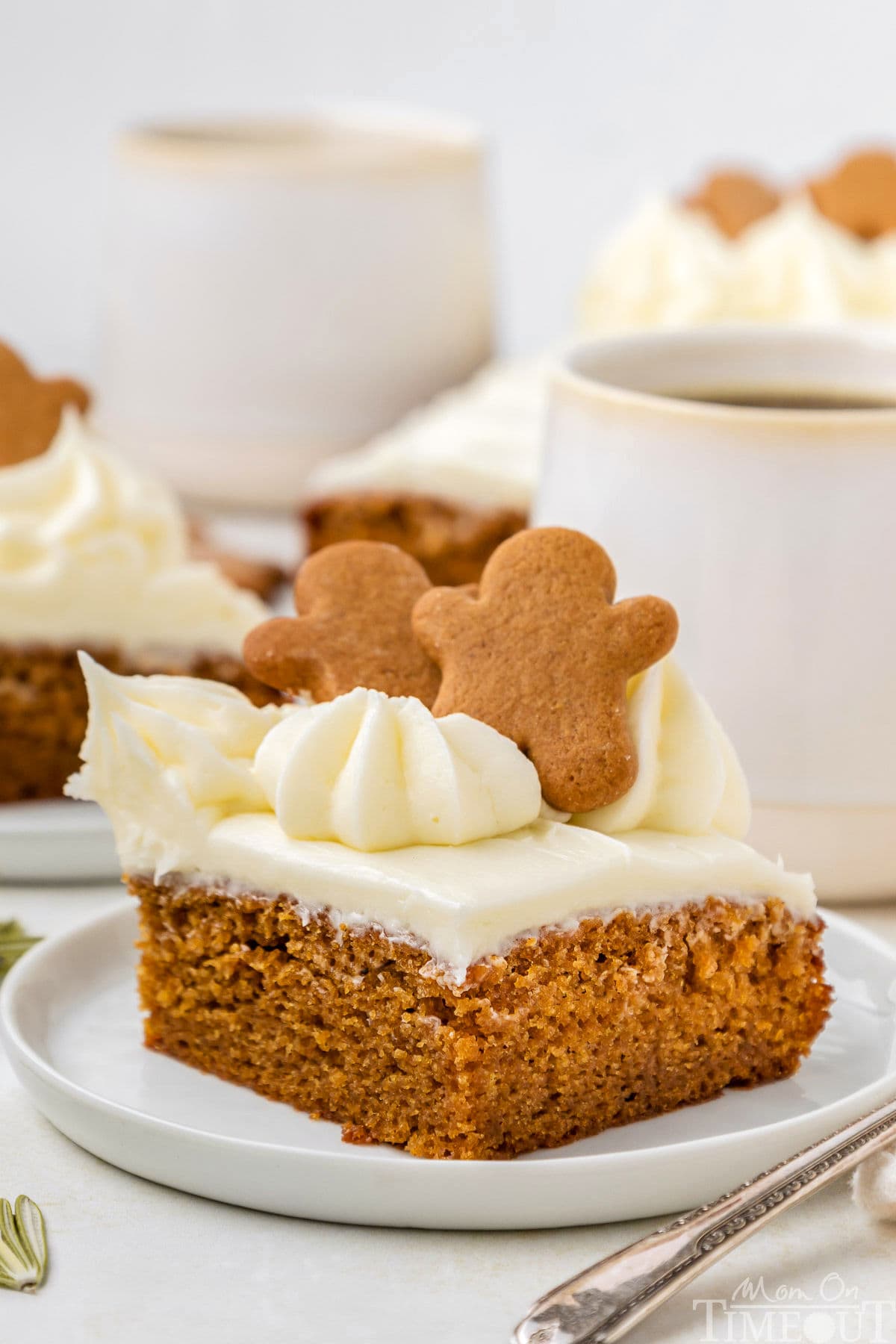 A slice of gingerbread cake topped with gingerbread men. The cake is on a white plate with mugs of coffee and more slices of cake in the background.