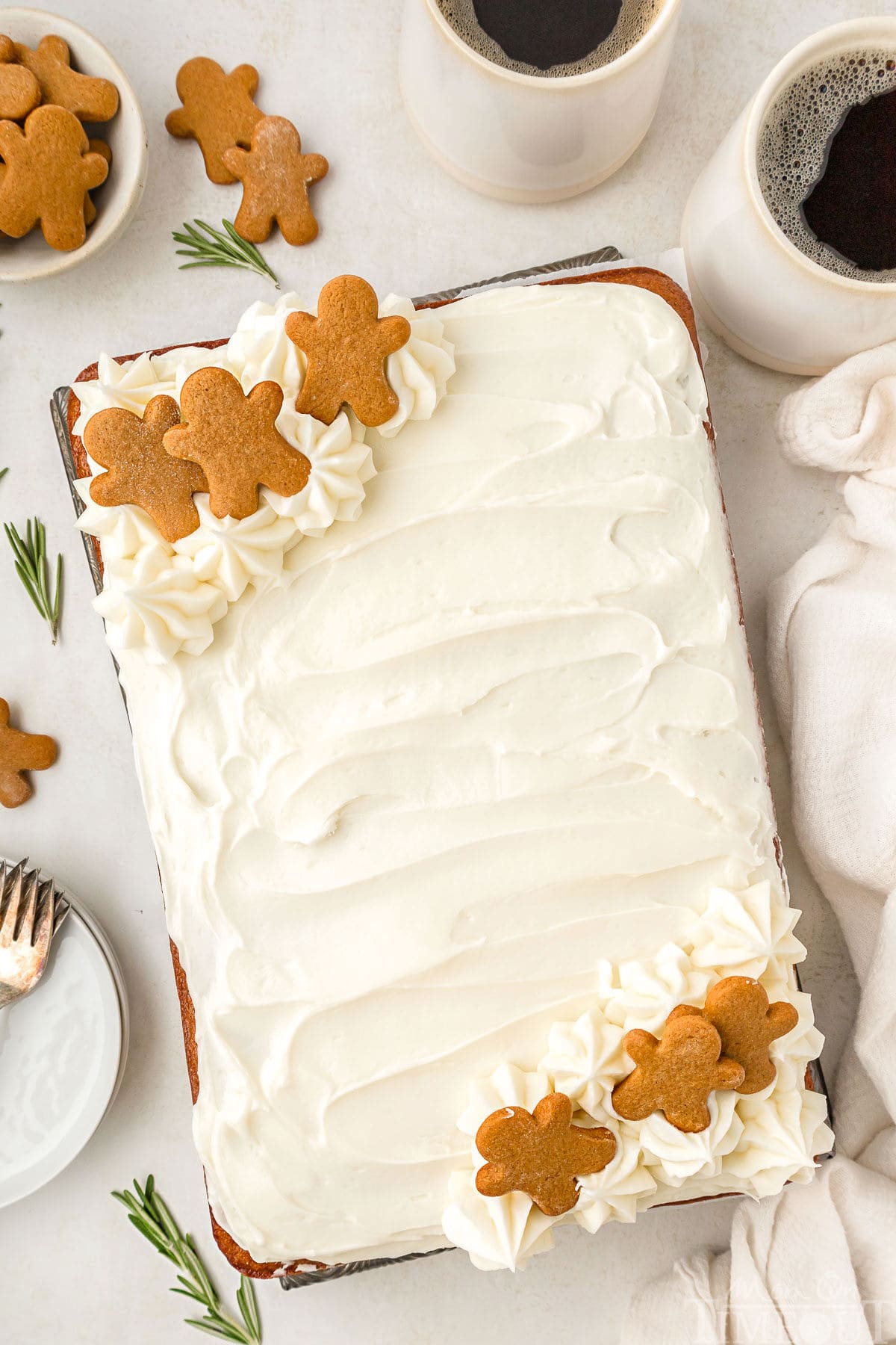 A top down view of gingerbread cake on a white surface. Mugs of coffee, gingerbread men, rosemary sprigs, plates, and a white linen are next to the ckae.