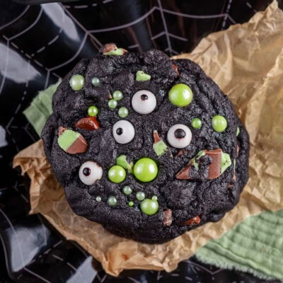 Monster Halloween Cookies stacked on brown parchment and black web design plate. Cookies have candy eyes and green sugar pearls for decoration.