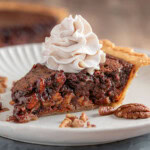 Generous slice of chocolate bourbon pecan pie on white plate topped with whipped cream. The pie can be seen in the background.