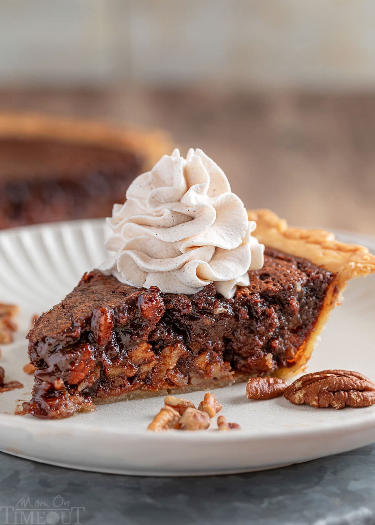 Generous slice of chocolate bourbon pecan pie on white plate topped with whipped cream. The pie can be seen in the background.