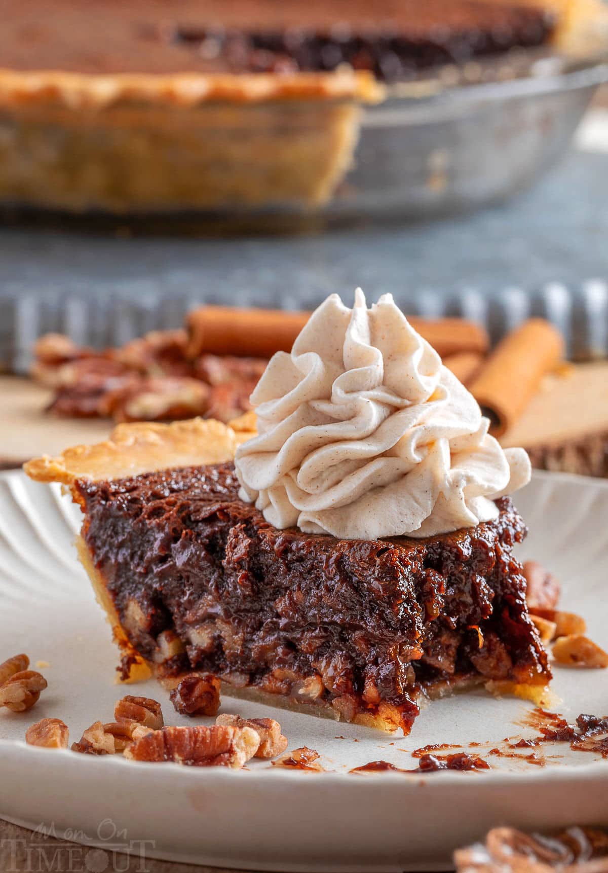 Generous slice of chocolate bourbon pecan pie on white plate topped with whipped cream. Bite has been taken from the end. The pie can be seen in the background.