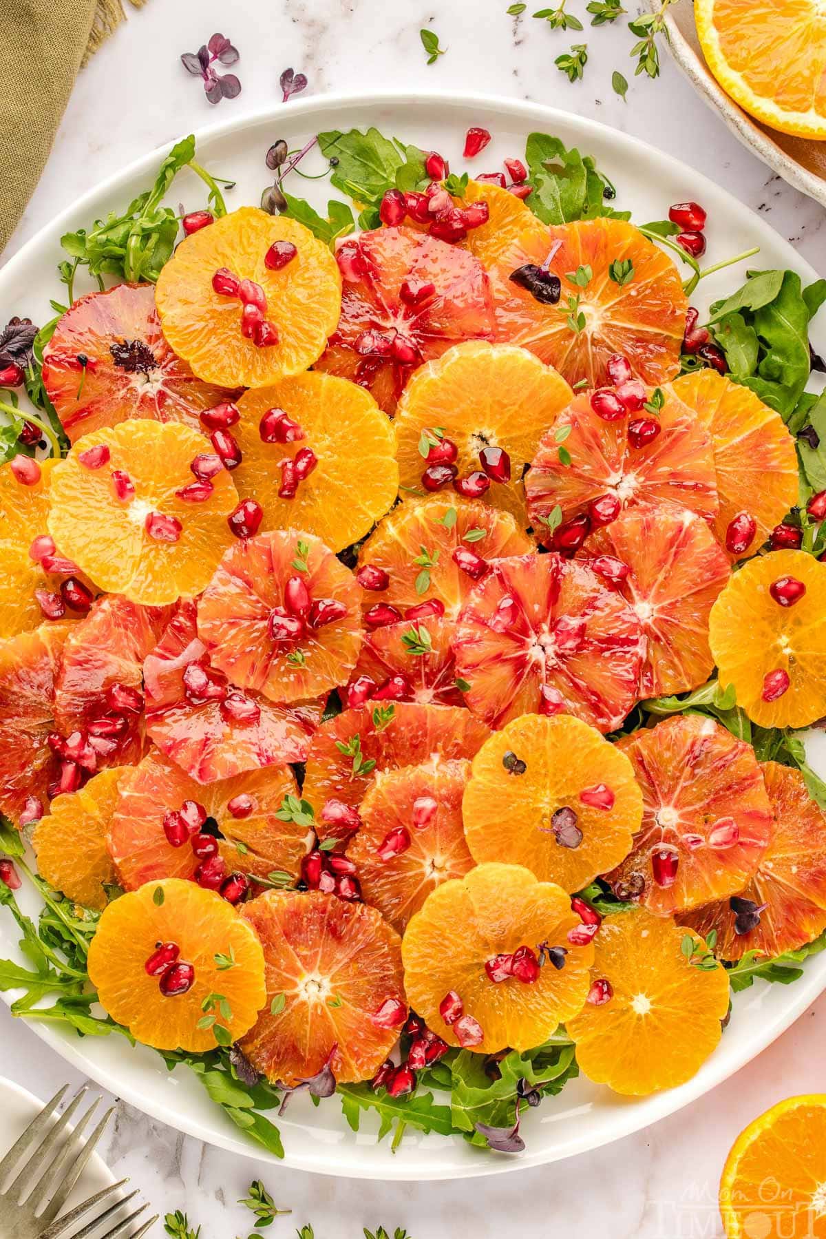 Close up look at citrus salad on white round platter ready to be served. Pomegranate seeds top the citrus rounds and arugula base.