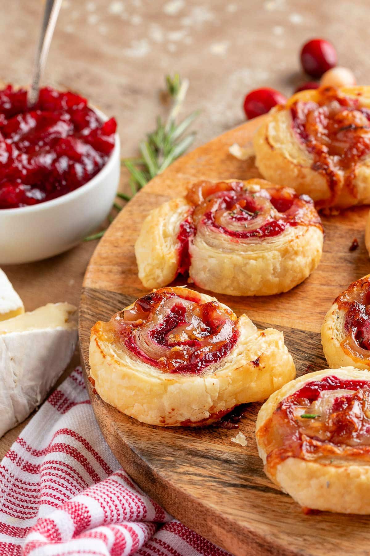 Cranberry brie puff pastry pinwheels on wood serving board. Cranberry sauce can be seen in the background.