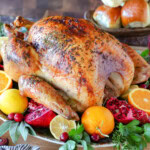 How To Cook A Turkey recipe with the whole roast turkey on a serving platter garnished with fresh fruit and herbs. Dinner rolls can be seen in the background.