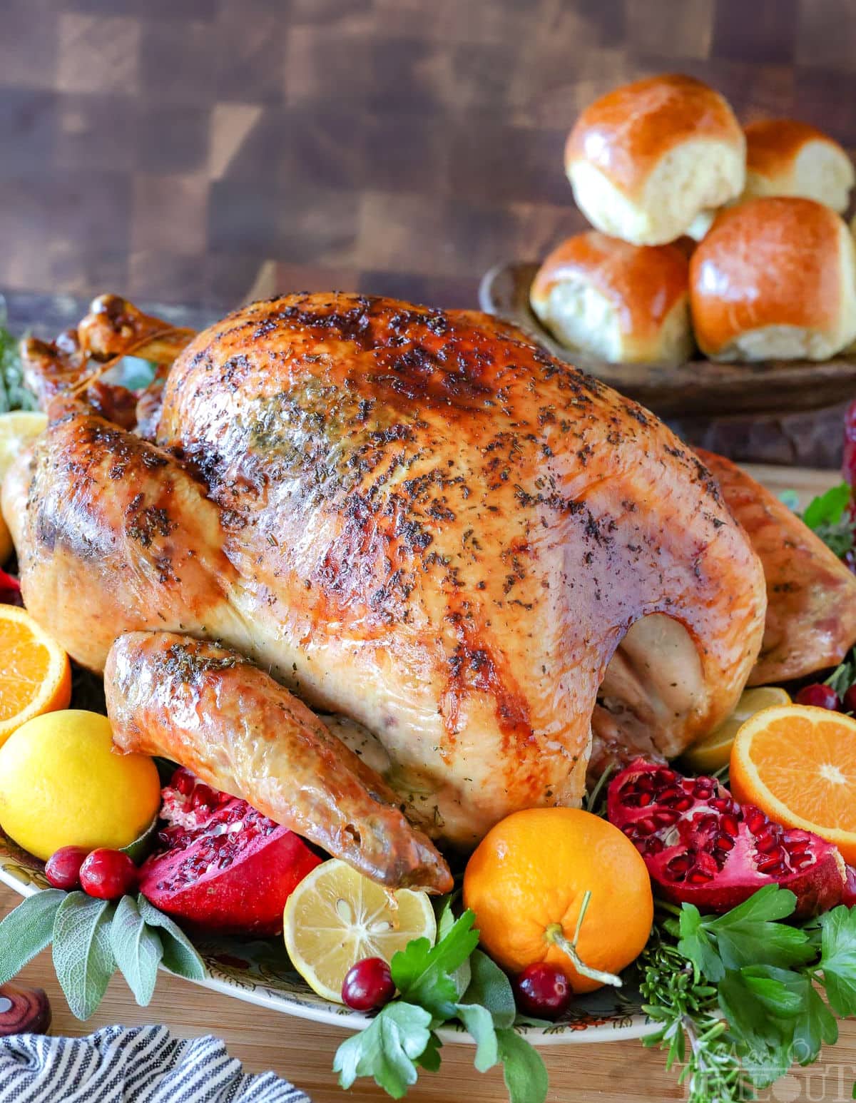 How To Cook A Turkey recipe with the whole roast turkey on a serving platter garnished with fresh fruit and herbs. Dinner rolls can be seen in the background.