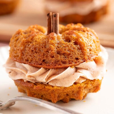 Mini pumpkin bundt cake made with two small bundts and sandwiched with cinnamon brown butter frosting. The stem is a cinnamon stick. The cake is sitting on a white plate with a fork.