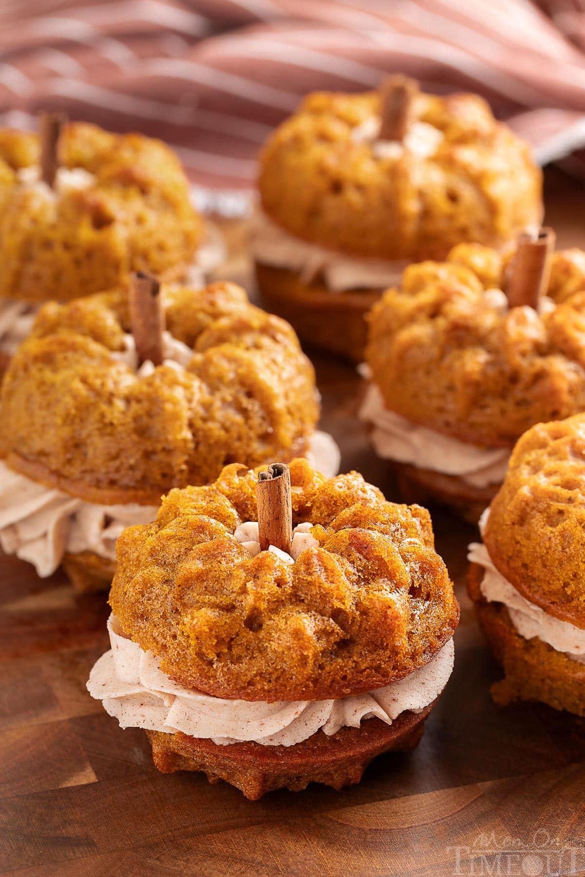 Six pumpkin bundt cakes with cinnamon stick stems sitting on dark wood board with striped towel in the background.