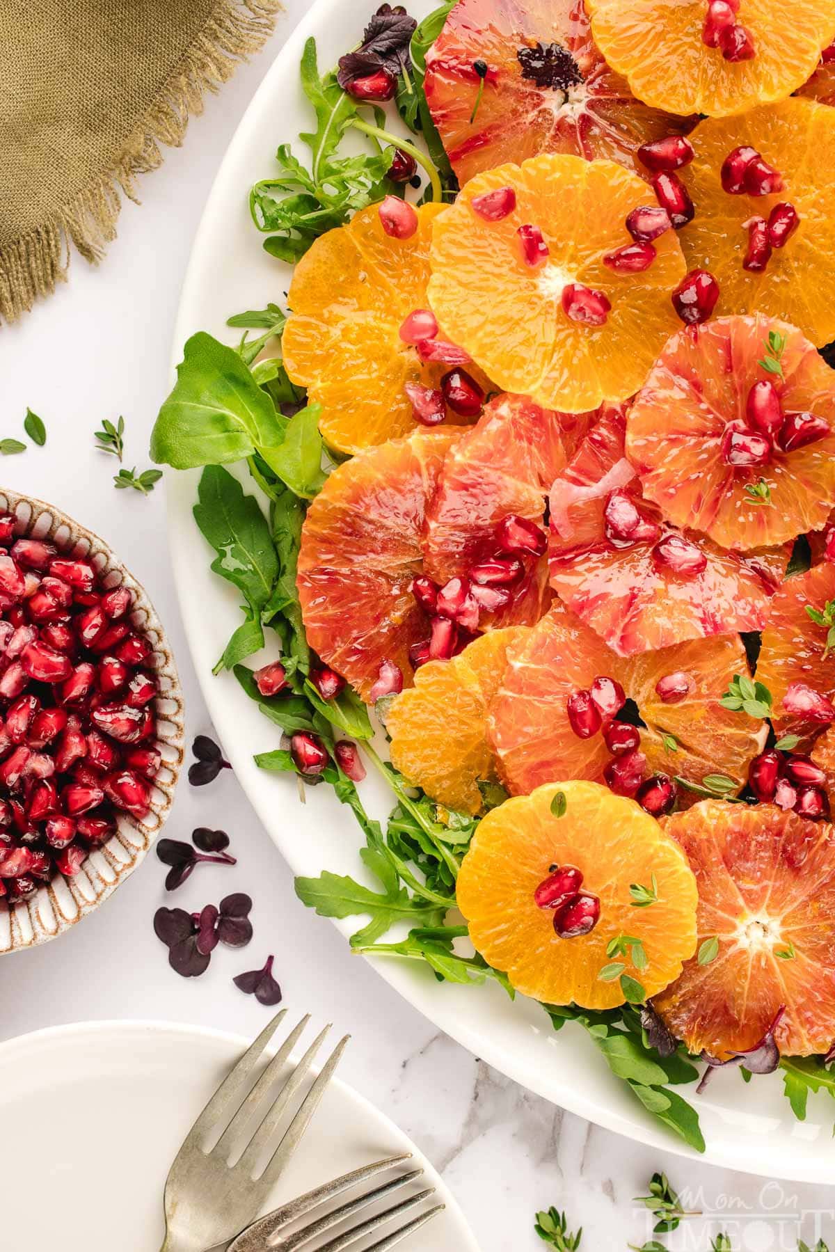 Winter citrus salad elegantly plated on large white platter and topped with thyme and pomegranate seeds. Oranges are cut in half and scattered around the white backdrop.