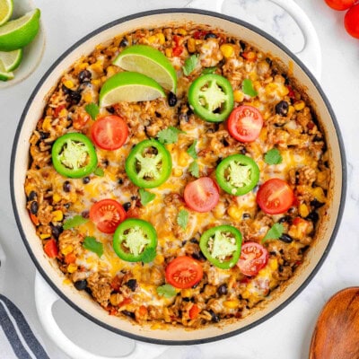 Top down view of taco rice in a white skillet topped with jalapeno slices, lime wedges and tomatoes. More taco topping can be seen scattered around the edge of the skillet.