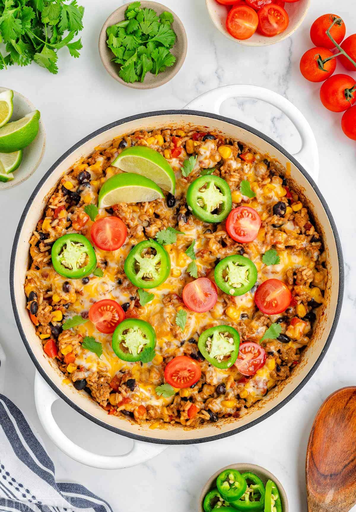 Top down view of taco rice in a white skillet topped with jalapeno slices, lime wedges and tomatoes. More taco topping can be seen scattered around the edge of the skillet.