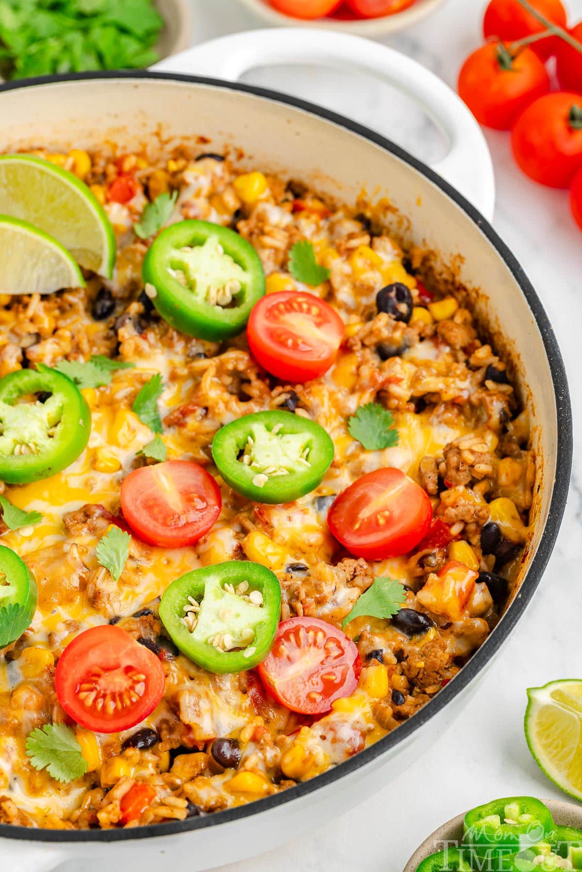 Close up view of taco rice in a white skillet topped with jalapeno slices, lime wedges and tomatoes. More taco topping can be seen scattered around the edge of the skillet.