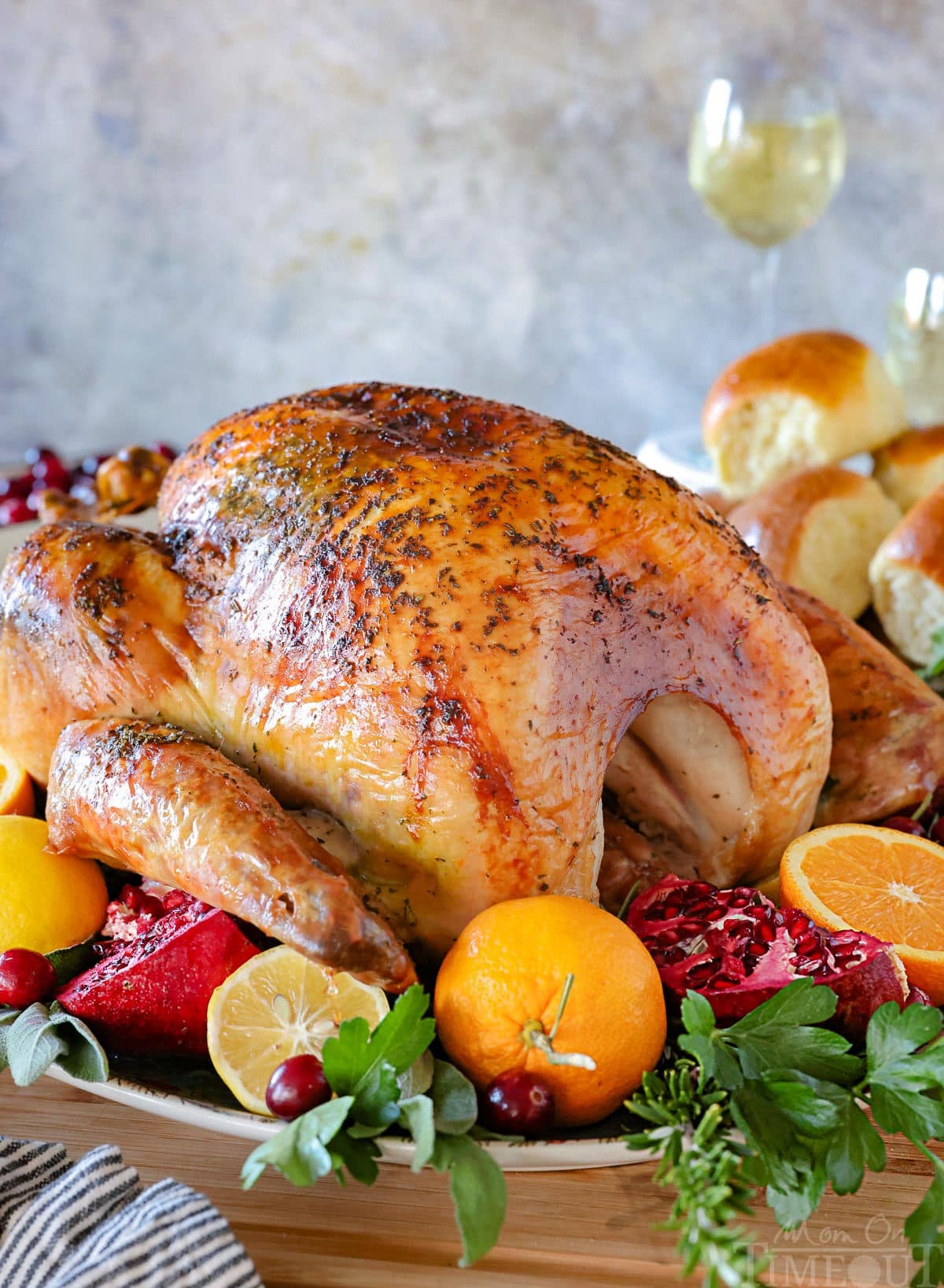 A perfect turkey recipe on a large serving platter roasted until golden brown and surround by citrus, herbs and cranberries. Dinner rolls can be seen in the background.