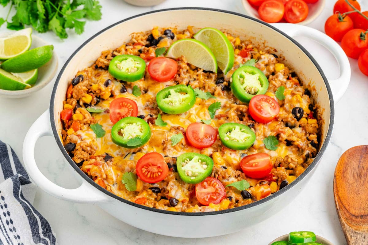 Top down angled view of taco rice in a white skillet topped with jalapeno slices, lime wedges and tomatoes. More taco topping can be seen scattered around the edge of the skillet.