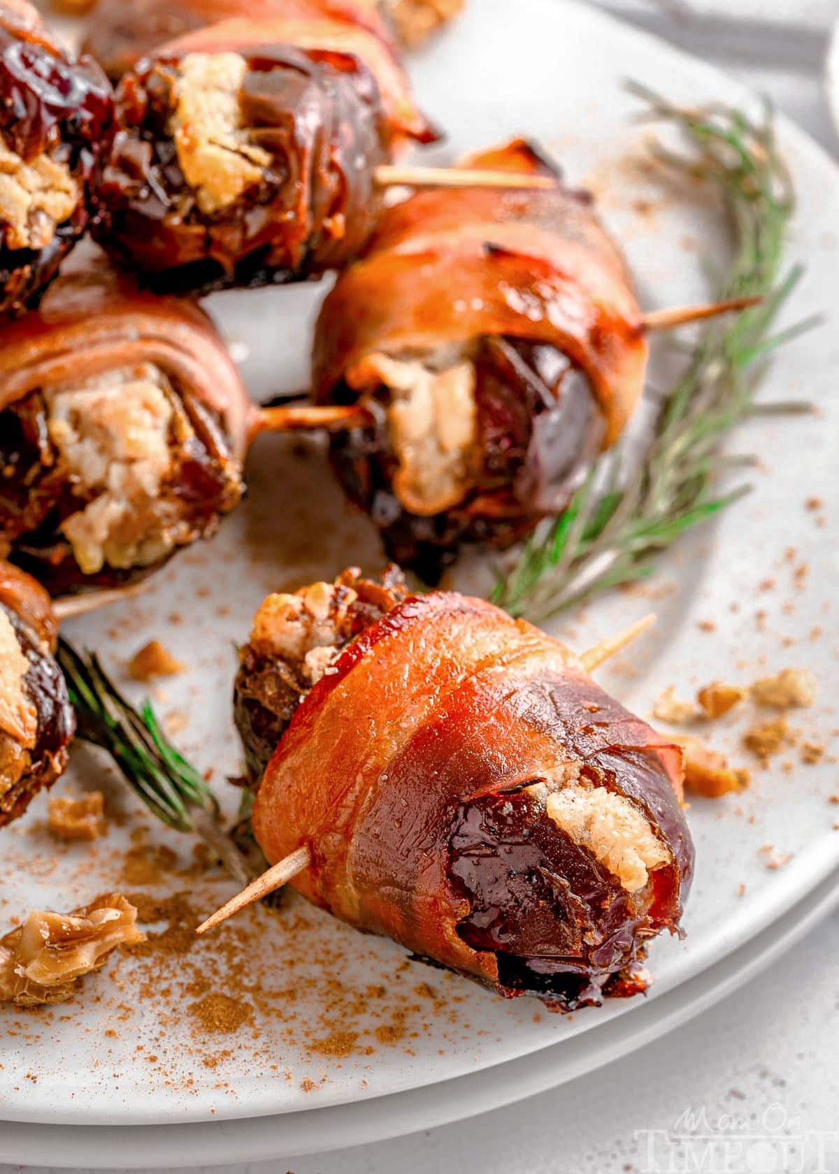 Shot of dates wrapped in bacon served on round white plate garnished with rosemary.