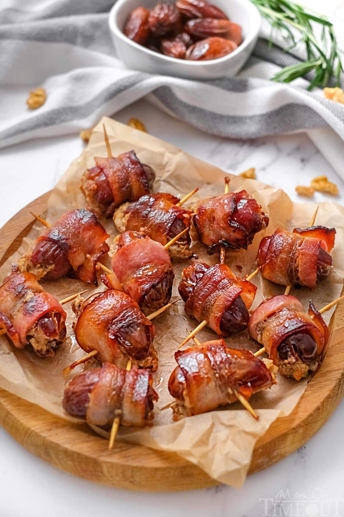 Wood serving tray topped with brown parchment paper and bacon wrapped dates ready to be enjoyed. The dates still have the toothpicks in them making them easy to pick up.