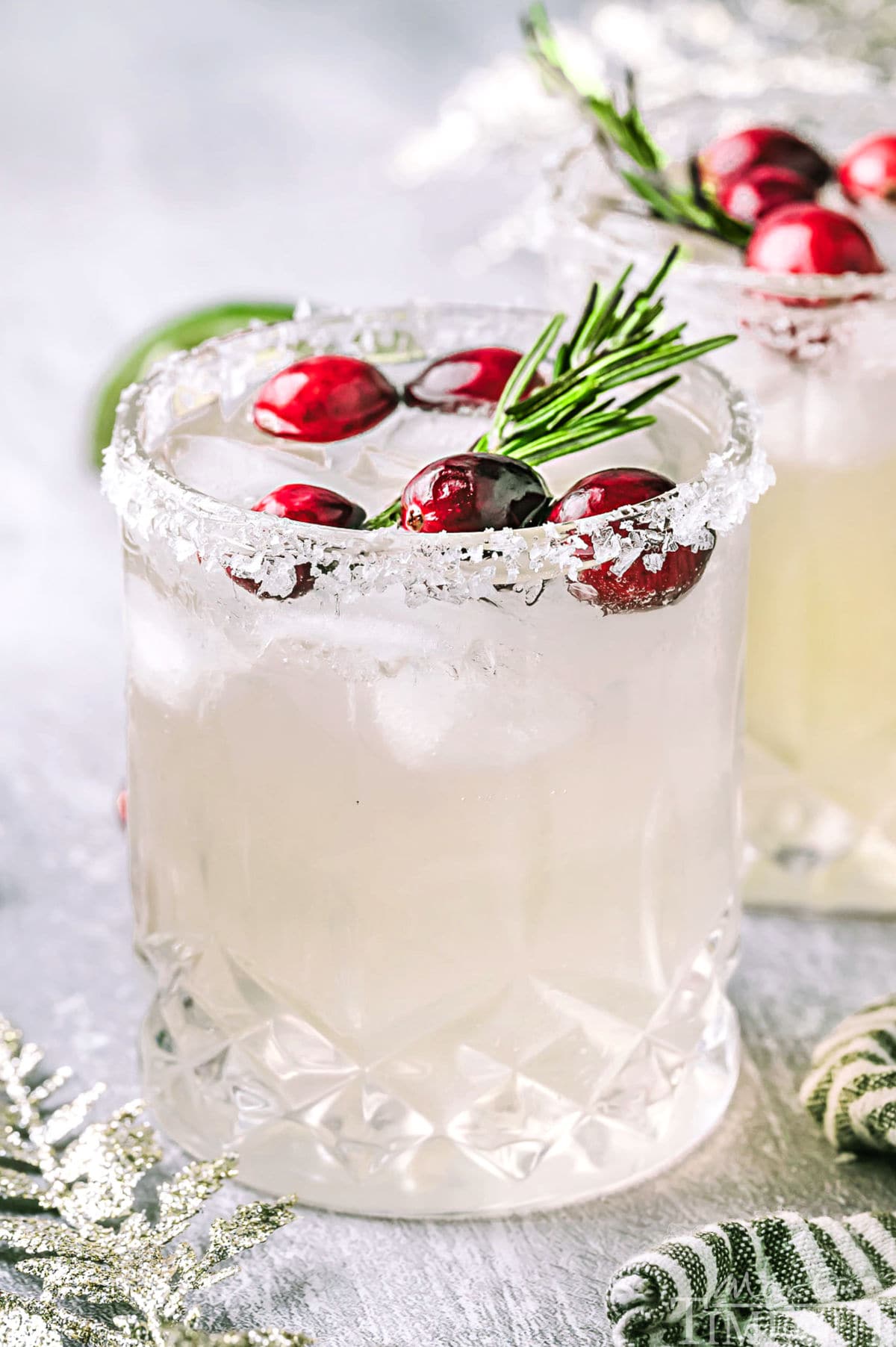 Christmas margarita in a short glass topped with fresh cranberries and a sprig of rosemary, Another glass can be seen in the background.