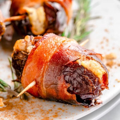 Shot of dates wrapped in bacon served on round white plate garnished with rosemary.